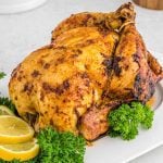 A whole rotisserie chicken with golden, crispy skin is served on a white platter, garnished with fresh parsley and lemon slices.