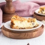 A slice of Chocolate Chip Cookie Cheesecake with a crumbly crust and chocolate chips sits on a white plate atop a round wooden tray, with a blurred background featuring a pink cloth and another slice of cake.