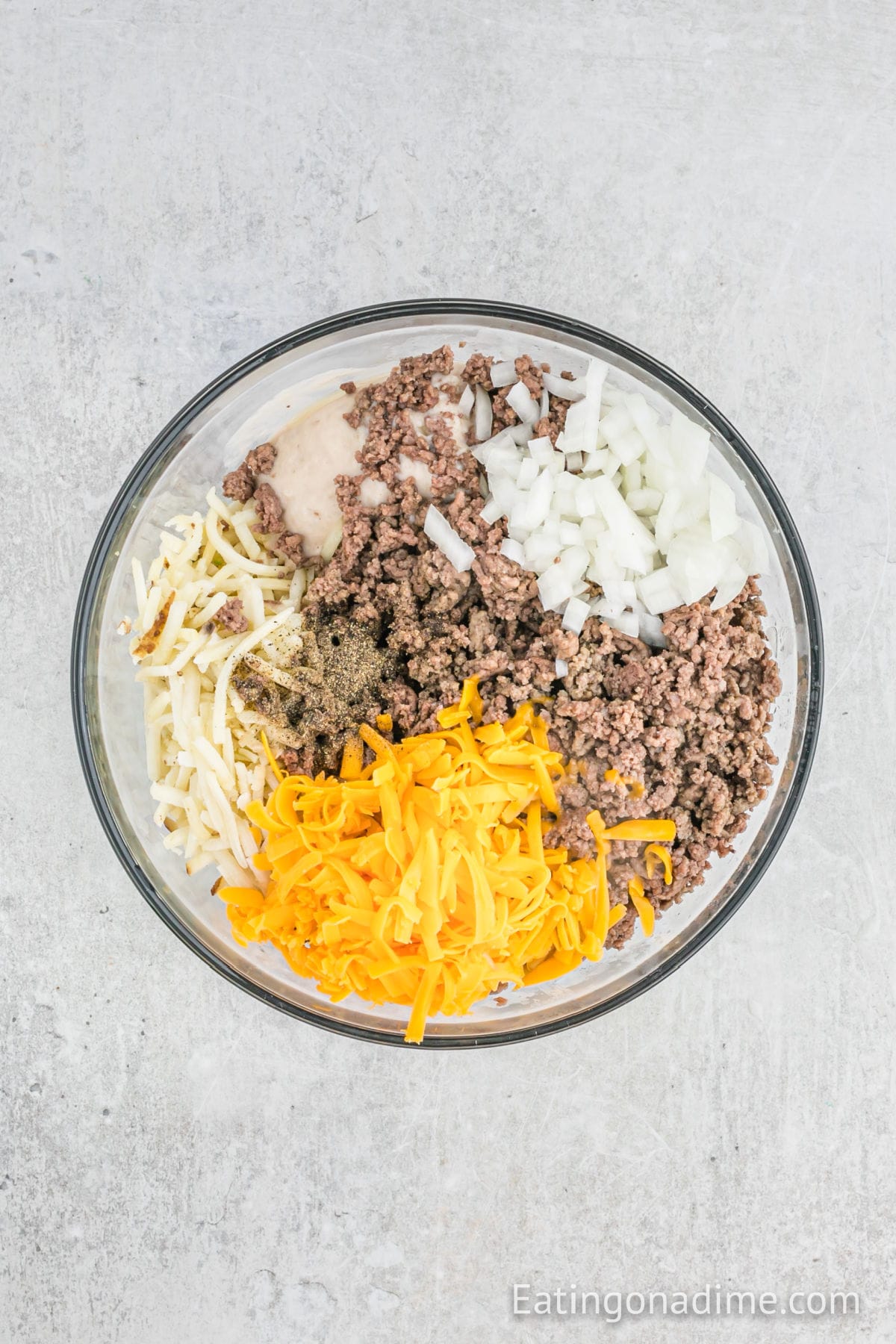 A glass bowl on a light surface contains cooked ground beef, chopped onions, shredded yellow and white cheese, seasonings, and creamy sauce—classic ingredients for Hamburger Hashbrown Casserole—arranged in separate sections.