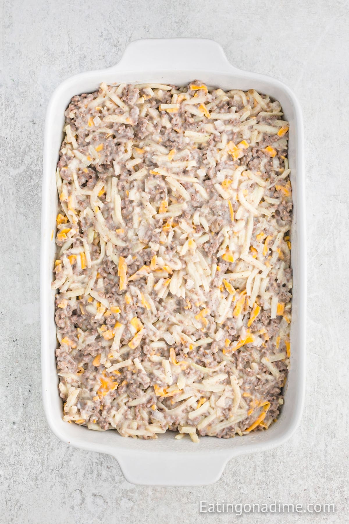 A white casserole dish filled with a creamy, well-mixed Hamburger Hashbrown Casserole—shredded potatoes, ground meat, and melted cheese layered together and ready to be baked.