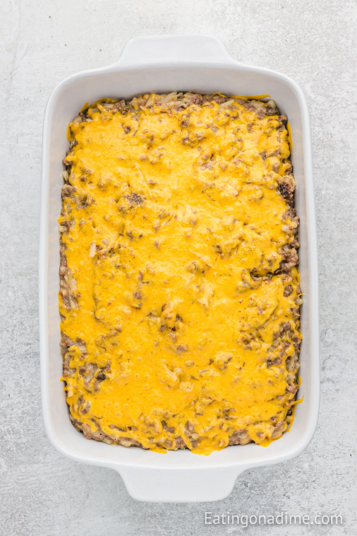 A white baking dish filled with Hamburger Hashbrown Casserole, topped with melted cheddar cheese, sits on a light gray surface.