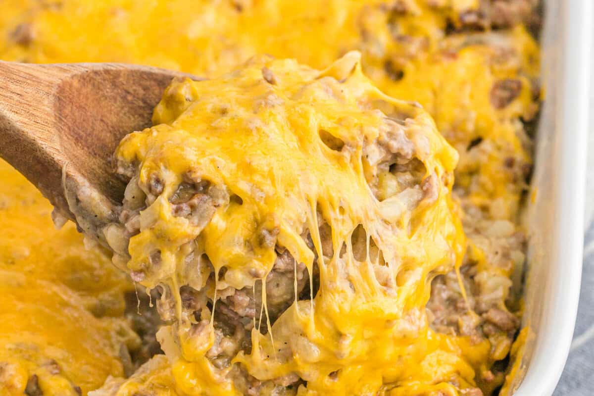 A wooden spoon scoops up a serving of Hamburger Hashbrown Casserole with ground beef and melted cheddar cheese, showing gooey, stretchy cheese on top.