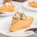 A slice of creamy No Bake Pumpkin Pie topped with whipped cream and a sprinkle of cinnamon, served on a white plate with a fork. Other slices are visible in the background.