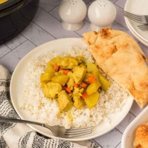 A plate of white rice topped with slow cooker chicken curry featuring chunks of chicken, carrots, and potatoes, served with a slice of naan bread. A fork, checkered cloth, and salt and pepper shakers are nearby on the table.