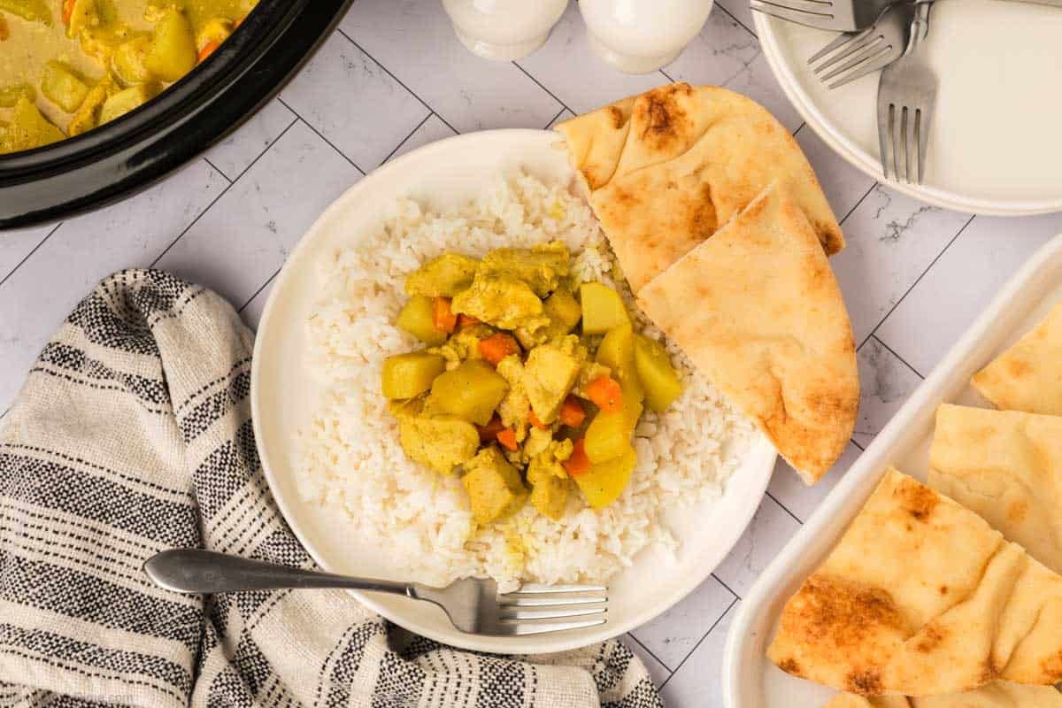 A plate of white rice topped with flavorful Slow Cooker Chicken Curry Recipe, filled with chicken and vegetables, served with slices of naan bread. A fork rests on the plate, a striped towel is nearby, and extra naan is on a separate dish.
