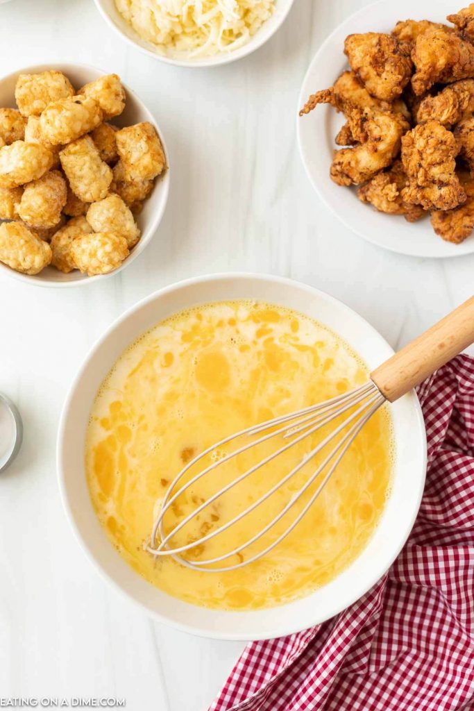 Whisking eggs in a white bowl with a bowl of tator tots, cheese and chicken nuggets
