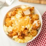 A Chick-Fil-A Breakfast Bowl features a white bowl filled with scrambled eggs, crispy fried chicken pieces, and shredded cheddar cheese, set on a white surface beside a red and white checkered cloth.