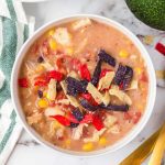 A bowl of creamy Chick-Fil-A Chicken Tortilla Soup topped with colorful tortilla strips, featuring visible corn, black beans, tomatoes, and chicken pieces. The bowl is placed on a marble surface next to a striped napkin.