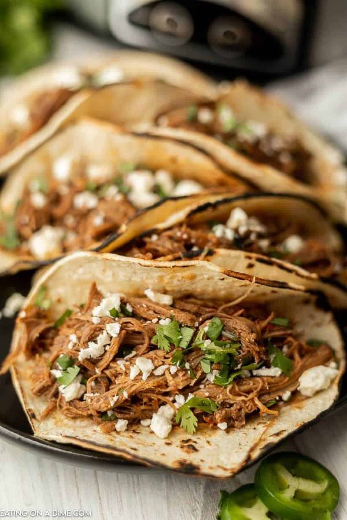 Three corn tortillas filled with slow cooker brisket, garnished with chopped cilantro and crumbled cheese. Slices of jalapeño are visible in the foreground, and a blurry background features a kitchen appliance.