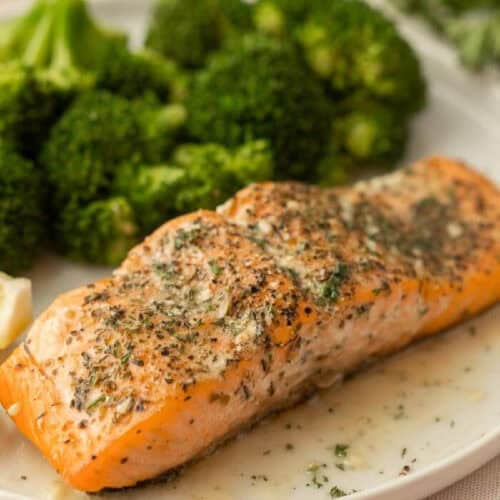 A seasoned baked salmon fillet, reminiscent of Olive Garden's Herb Grilled Salmon, is garnished with herbs and served on a plate alongside fresh, steamed broccoli. A lemon wedge is partially visible on the side, enhancing the dish's presentation.
