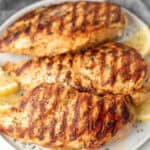 Three grilled chicken breasts with char marks are served on a white plate, infused with the best chicken marinade, garnished with chopped herbs, and accompanied by lemon wedges.