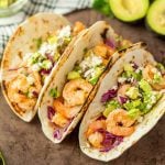Three Grilled Shrimp Tacos on grilled tortillas, filled with cooked shrimp, chopped avocado, shredded red cabbage, diced onions, cilantro, and crumbled cheese, are arranged on a brown surface with avocado halves in the background.