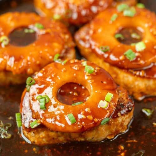 Close-up of Hawaiian pork chops in a skillet. The pork chops are topped with pineapple slices and garnished with chopped green onions, all coated in a thick, rich sauce.