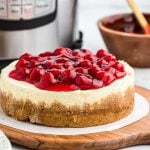An Instant Pot Cheesecake with a graham cracker crust is topped with glossy cherry pie filling, sitting on round parchment paper on a wooden board. An Instant Pot and a bowl of extra cherry topping are in the background.