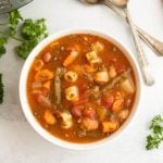 A bowl of vegetable soup inspired by instant pot minestrone soup recipes, filled with pasta, beans, carrots, green beans, and potatoes in a tomato broth, garnished with herbs. Two spoons and fresh parsley surround the bowl on a white surface.