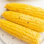 Three ears of cooked corn on the cob, lightly seasoned with black pepper, are arranged on a white plate.