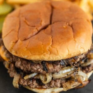 A close-up of a double cheeseburger on a soft bun, featuring juicy beef patties and grilled onions. Pickles and French fries are slightly blurred in the background.