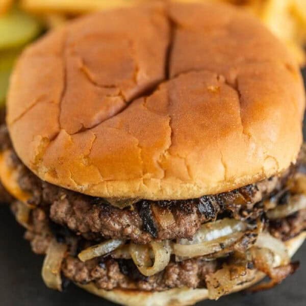 A close-up of a double cheeseburger on a soft bun, featuring juicy beef patties and grilled onions. Pickles and French fries are slightly blurred in the background.