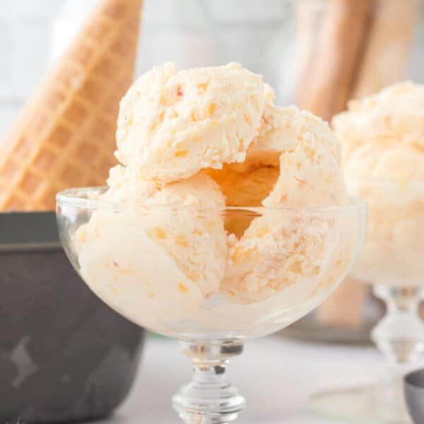 A glass dish holds several scoops of light-colored homemade peach ice cream, capturing the essence of summer. A waffle cone is visible in the background, blurred slightly. The setting appears to be a bright kitchen or dining area.