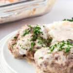 Two creamy Swedish meatballs, inspired by Poor Man's Steak, are garnished with chopped parsley on a white plate, served with mashed potatoes and gravy. A glass baking dish with more meatballs is visible in the background.