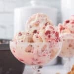 A close-up of a glass bowl filled with creamy swirls of red velvet ice cream in pink and white hues. The dessert looks tempting with visible chunks of flavor, set against a soft white and gray background.