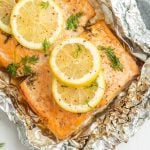 Baked salmon fillet in foil, reminiscent of grilled salmon in foil, topped with lemon slices and garnished with fresh dill. The crinkled foil wraps the fish as it bakes, lightly seasoned with pepper.