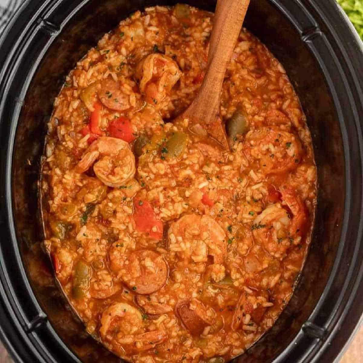 A crock pot shrimp jambalaya recipe is shown, with rice, sausage slices, shrimp, diced tomatoes, bell peppers, and spices. A wooden spoon stirs the hearty, colorful mixture in the slow cooker.