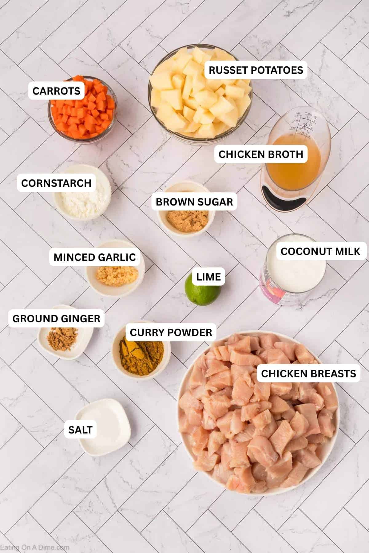 Ingredients for recipe: diced carrots, potatoes, chicken broth, milk, cubed raw chicken, lime, curry powder, minced garlic, brown sugar, flour, and salt on a white tiled surface.