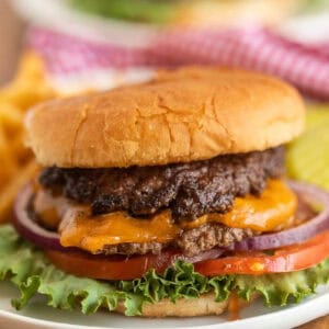 A close-up of a Smash Burger Recipe featuring melted cheddar cheese, lettuce, tomato, red onion, and a juicy beef patty in a soft bun, with crinkle-cut fries and pickles in the background.