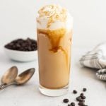 A tall glass of iced coffee, reminiscent of a Starbucks Caramel Frappuccino, topped with whipped cream and caramel drizzle. Coffee beans, two spoons, and a bowl of coffee beans rest in the background on a light surface.