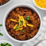 A bowl of Texas Roadhouse Chili topped with shredded cheddar cheese and chopped green onions, surrounded by a striped napkin, a spoon, and small bowls of extra cheese and green onions.