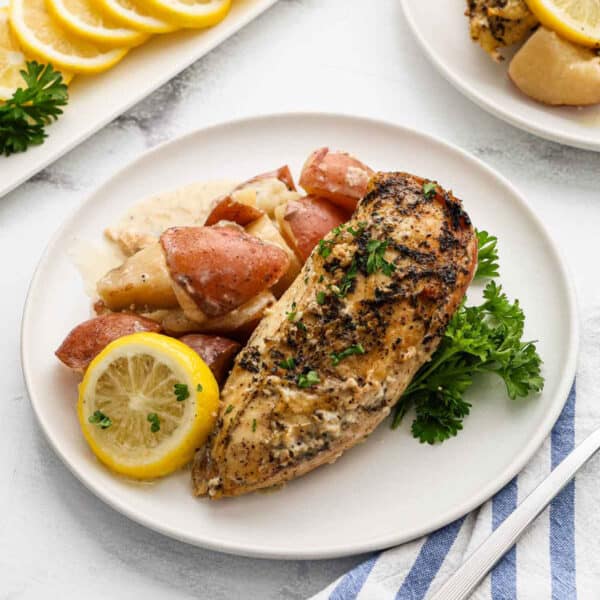 A plate with a seasoned baked chicken breast, reminiscent of a Slow Cooker lemon garlic chicken recipe, roasted red potatoes, a lemon slice, and parsley, set on a white table next to a blue-striped napkin.