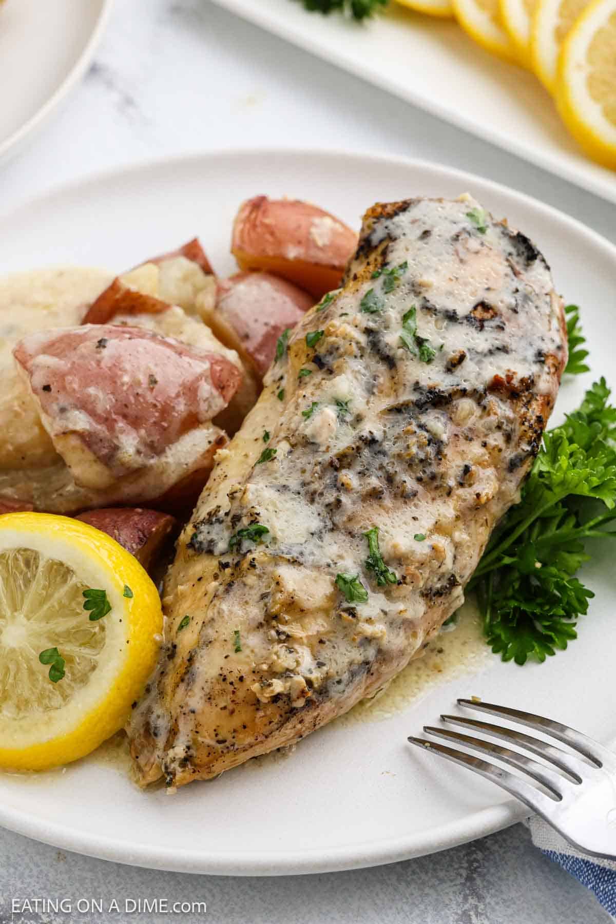 A plate with a grilled chicken breast topped with creamy sauce, garnished with parsley. Slices of lemon and roasted red potatoes are served on the side—try this as a twist on your favorite Slow Cooker lemon garlic chicken recipe.