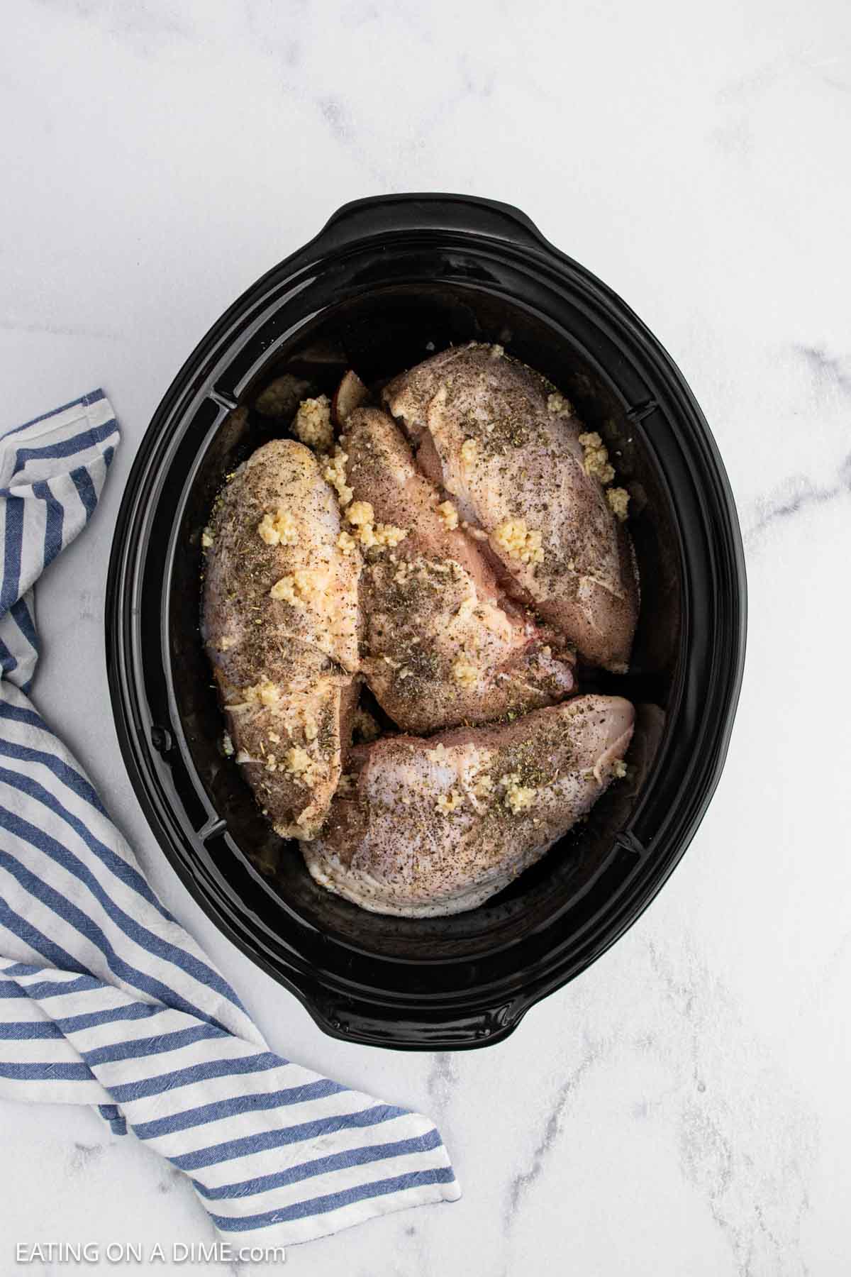Raw, seasoned chicken breasts topped with minced garlic are arranged in a black slow cooker for this Slow Cooker lemon garlic chicken recipe. A blue and white striped kitchen towel rests beside the slow cooker on a white marble surface.