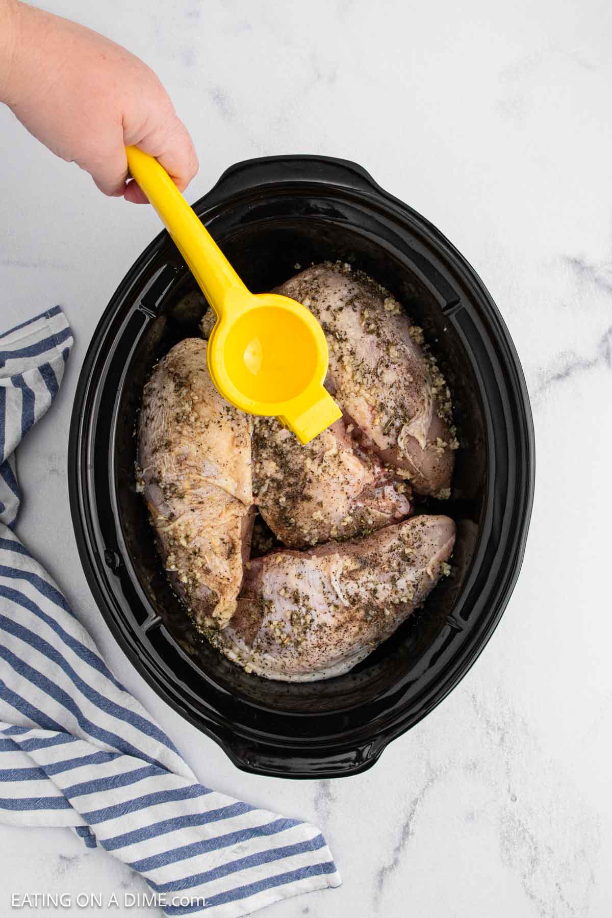 A hand squeezes a yellow lemon juicer over seasoned raw chicken breasts in a black slow cooker, perfect for a Slow Cooker lemon garlic chicken recipe, with a blue and white striped towel beside it on a marble surface.