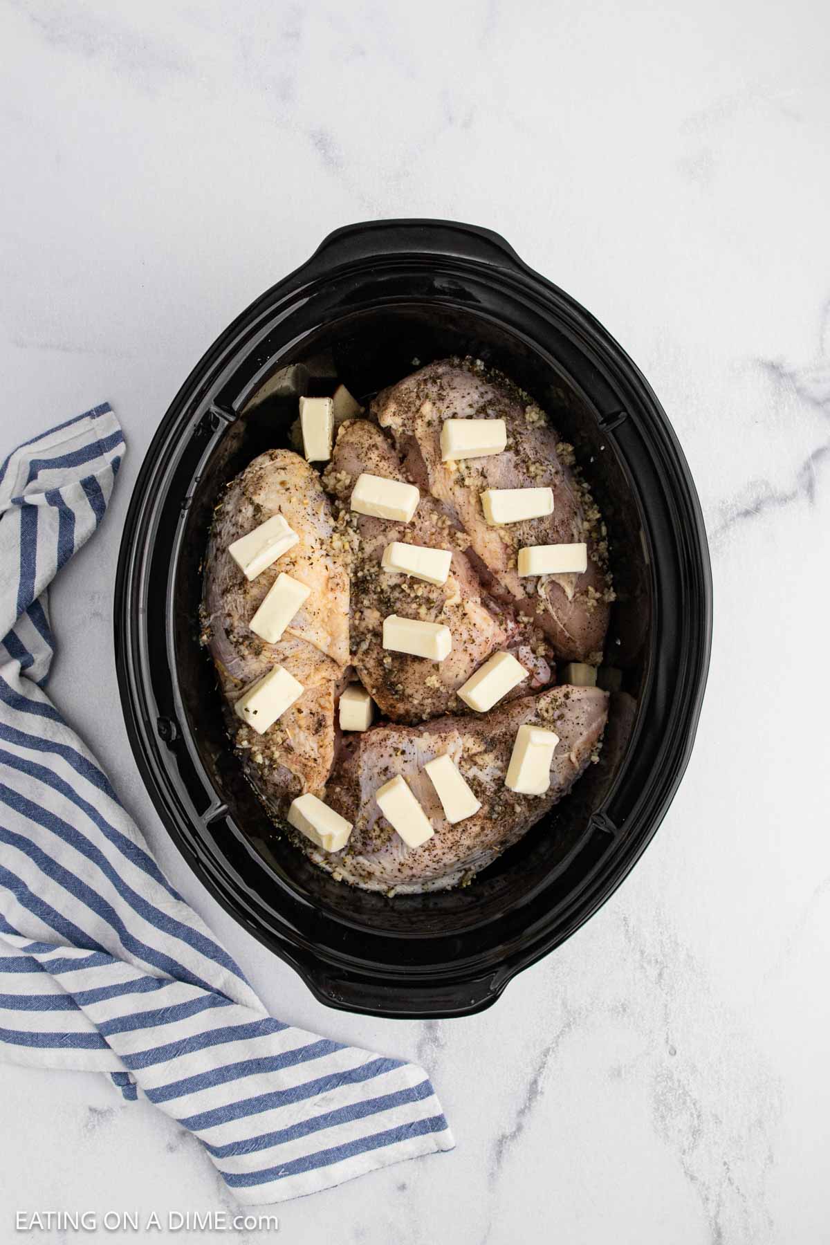 Chicken breasts topped with seasonings and cubed butter in a black slow cooker, perfect for a Slow Cooker lemon garlic chicken recipe, placed on a white marble surface next to a blue and white striped kitchen towel.