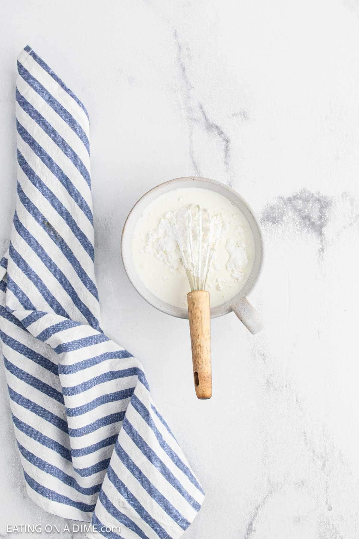 A bowl of white cream with a whisk inside sits on a light surface next to a blue and white striped cloth, ready for the Slow Cooker lemon garlic chicken recipe.