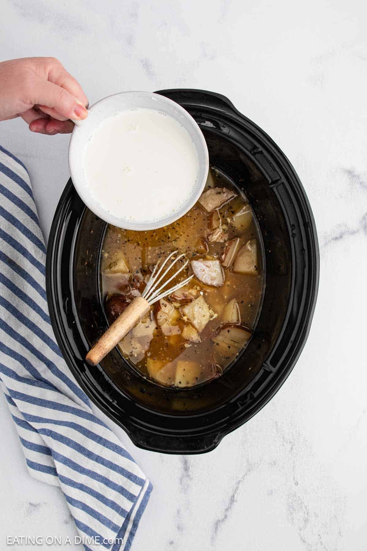 A hand pours a bowl of white liquid into a slow cooker filled with meat and broth for a Slow Cooker lemon garlic chicken recipe. A whisk rests inside the cooker, and a striped kitchen towel lies nearby on a marble surface.