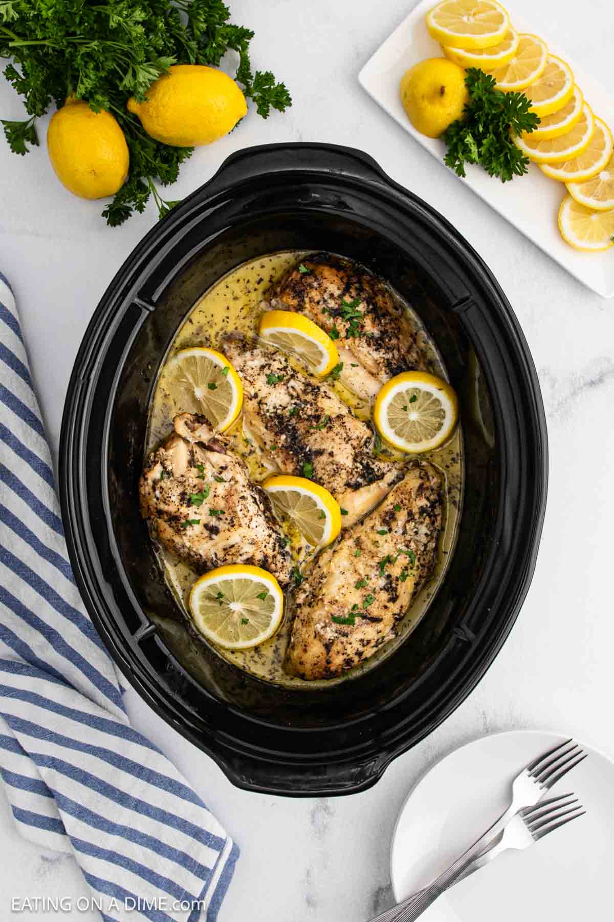 Slow Cooker lemon garlic chicken recipe featuring tender chicken breasts garnished with lemon slices and parsley. Fresh lemons, parsley, and a striped towel sit on a white countertop alongside two forks on a white plate.