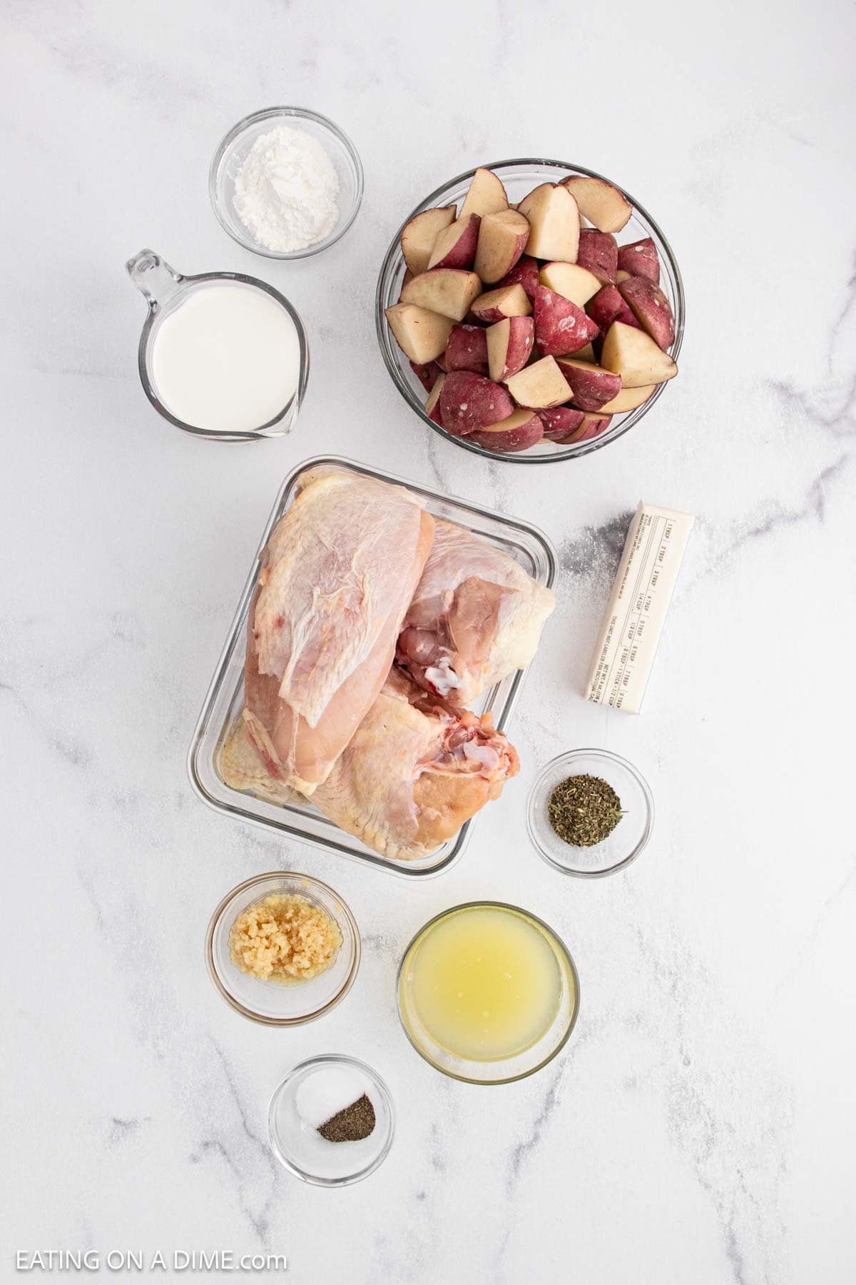 Top-down view of ingredients for a Slow Cooker lemon garlic chicken recipe on a white surface: raw chicken breasts, chopped red potatoes, butter, milk, flour, minced garlic, chicken broth, salt, pepper, and dried herbs in small bowls.