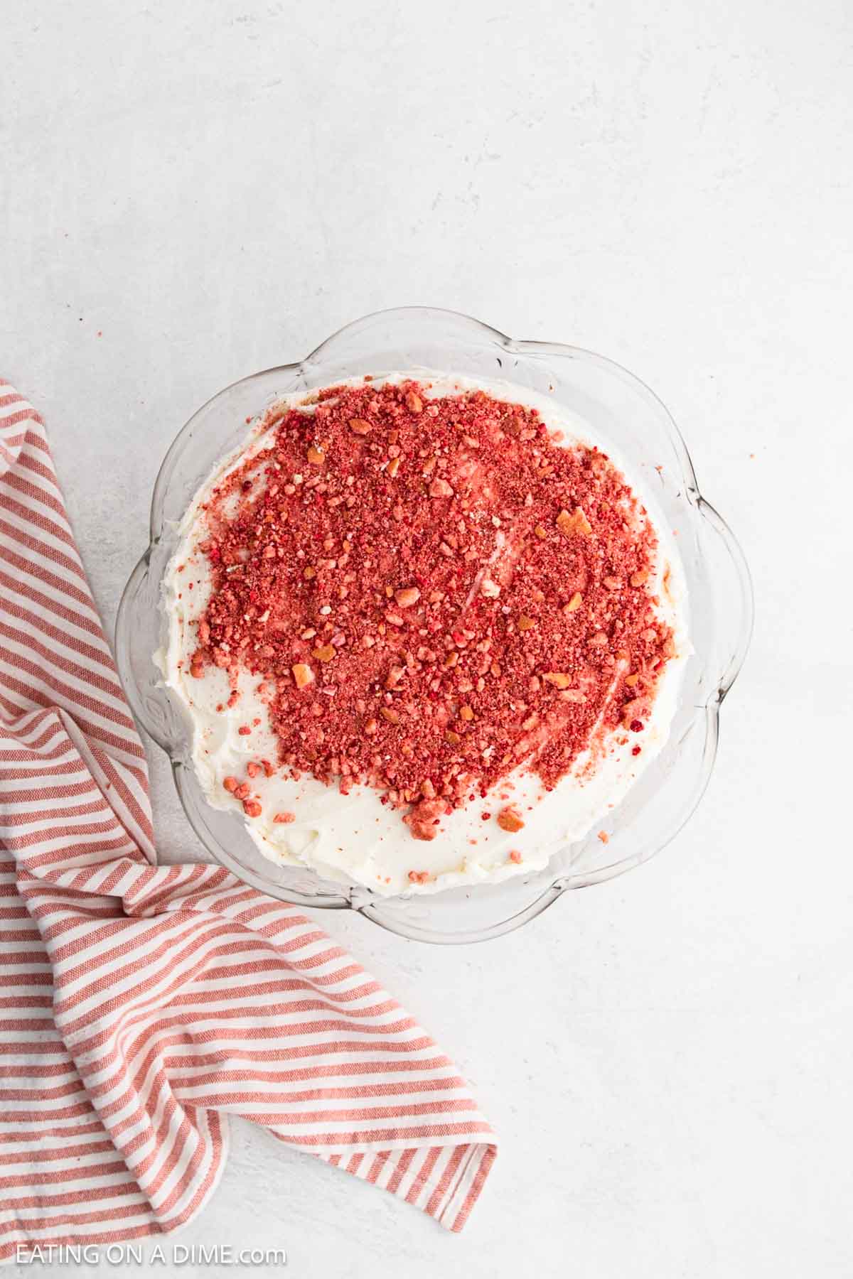A round Strawberry Crunch Cake with white frosting, topped with a generous layer of crumbled red topping, sits on a glass plate next to a pink and white striped cloth on a light surface.