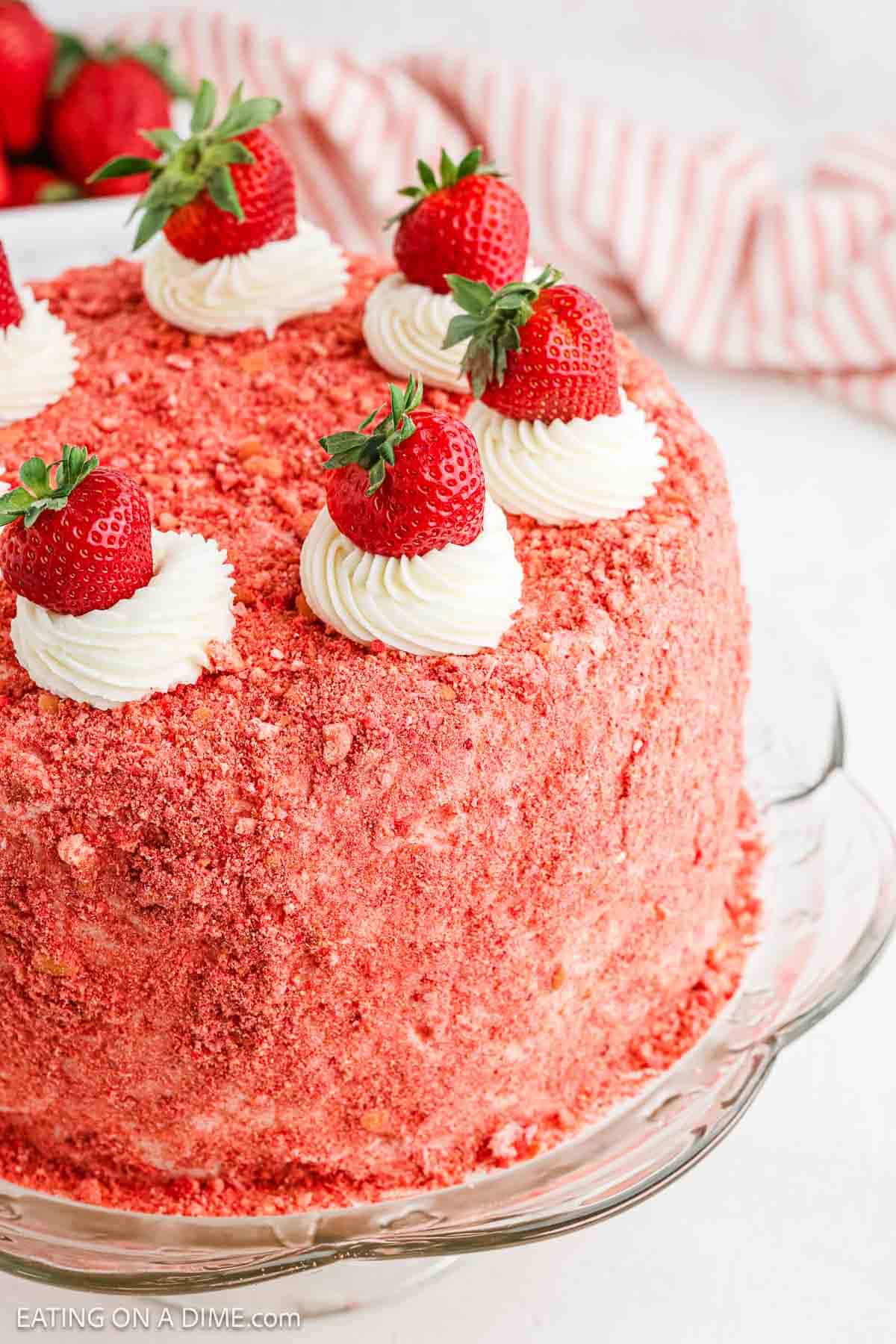 A Strawberry Crunch Cake with a crumbly pink coating, topped with swirls of white frosting and fresh strawberries, sits on a glass cake stand. A striped cloth is in the background.