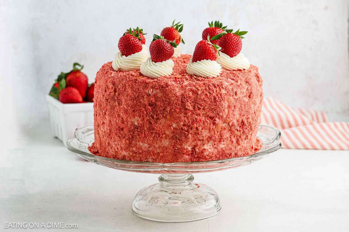 A round Strawberry Crunch Cake with a pink crumb coating, topped with swirls of white frosting and whole fresh strawberries, sits on a glass cake stand. A bowl of strawberries is in the background.