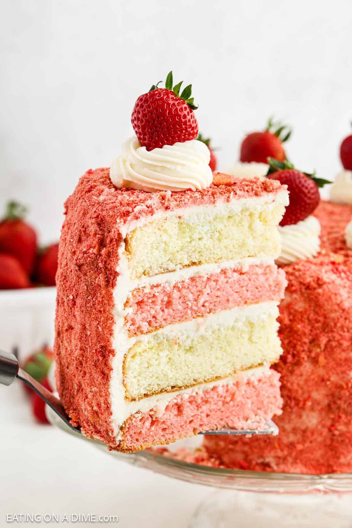 A slice of three-layer Strawberry Crunch Cake with pink and white layers, creamy white frosting, and a crumb coating. The slice is topped with whipped cream and a whole strawberry, with the entire cake visible in the background.