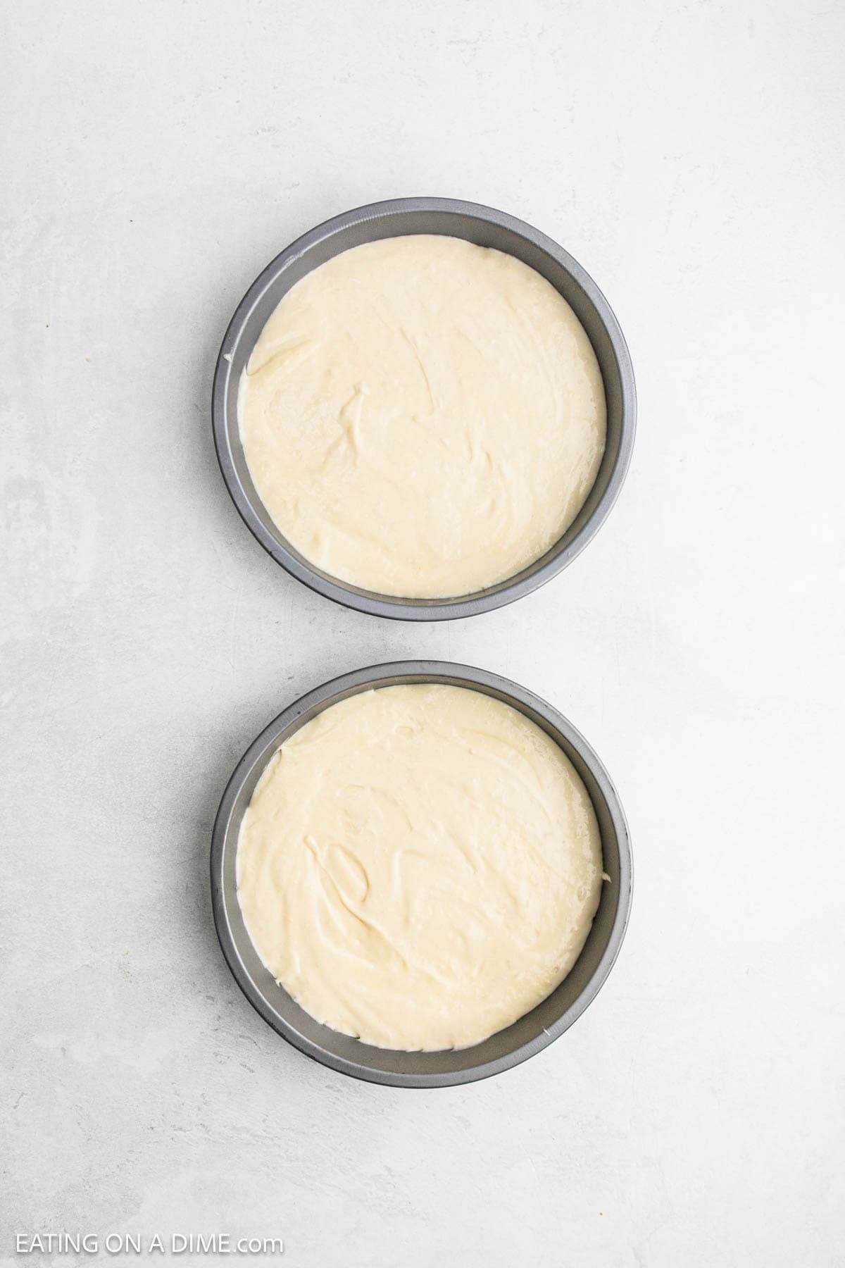 Two round cake pans filled with smooth, pale Strawberry Crunch Cake batter are placed on a light-colored surface, ready to be baked.