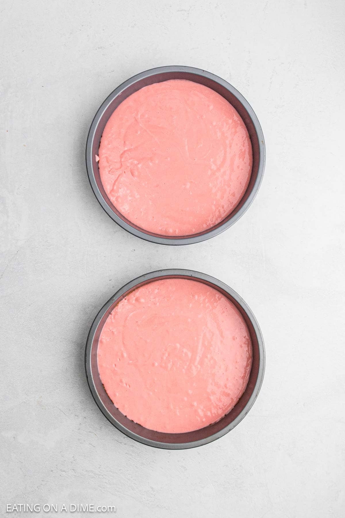 Two round cake pans filled with pink Strawberry Crunch Cake batter are placed on a light gray surface, ready to be baked.