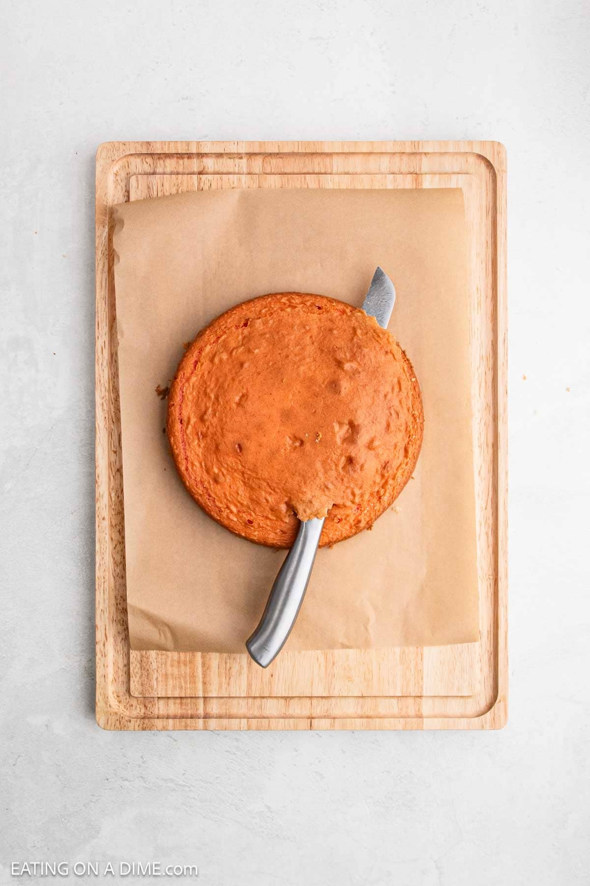 A round, golden-brown Strawberry Crunch Cake sits on parchment paper atop a wooden cutting board. A metal knife is partially inserted under the cake, ready to lift or cut it.