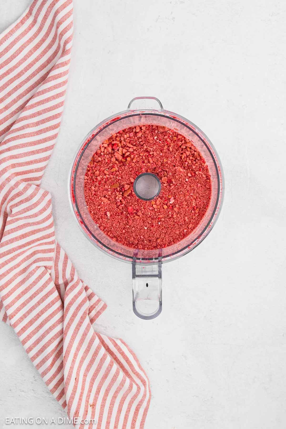 A food processor bowl filled with finely crushed red crumbs for a Strawberry Crunch Cake sits on a light surface next to a pink and white striped cloth.
