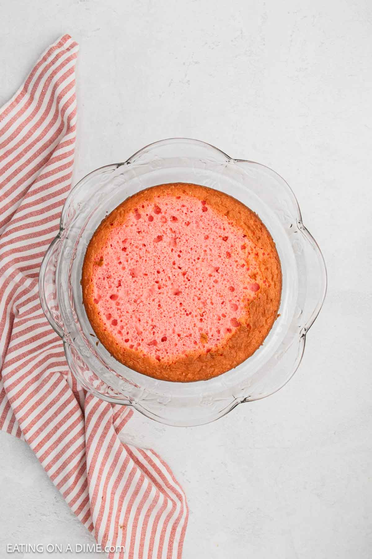 A round Strawberry Crunch Cake sits in a glass dish on a light surface, next to a red and white striped cloth.