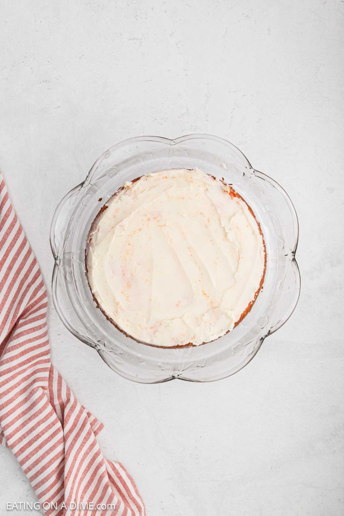 A round Strawberry Crunch Cake with white frosting sits on a clear glass plate. The background is a light, textured surface, and a pink and white striped cloth is placed to the left of the plate.